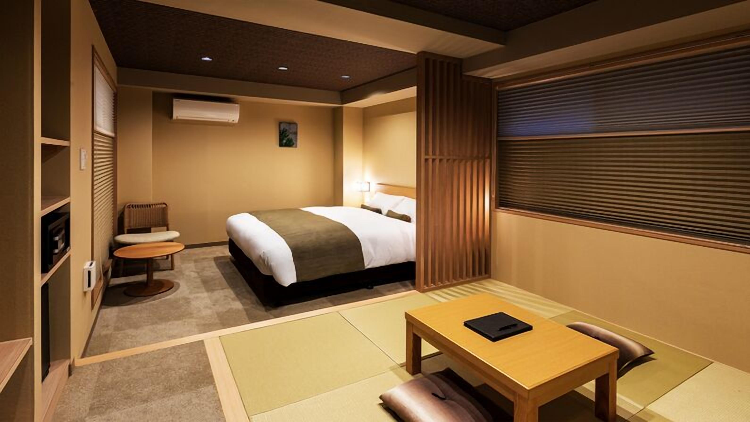 A hotel room with a bed, a desk, a chair, a radiator, and a window with blinds.