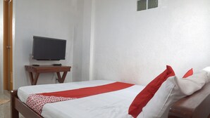 Una cama bien tendida con una colcha blanca y roja, una mesita de noche de madera con un televisor, y una pared blanca con una ventana pequeña.