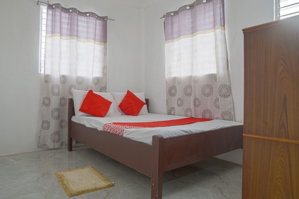 Una cama individual con almohadas rojas, cabecera de madera y colchón blanco. A la derecha hay un armario de madera y una alfombra pequeña en el suelo.