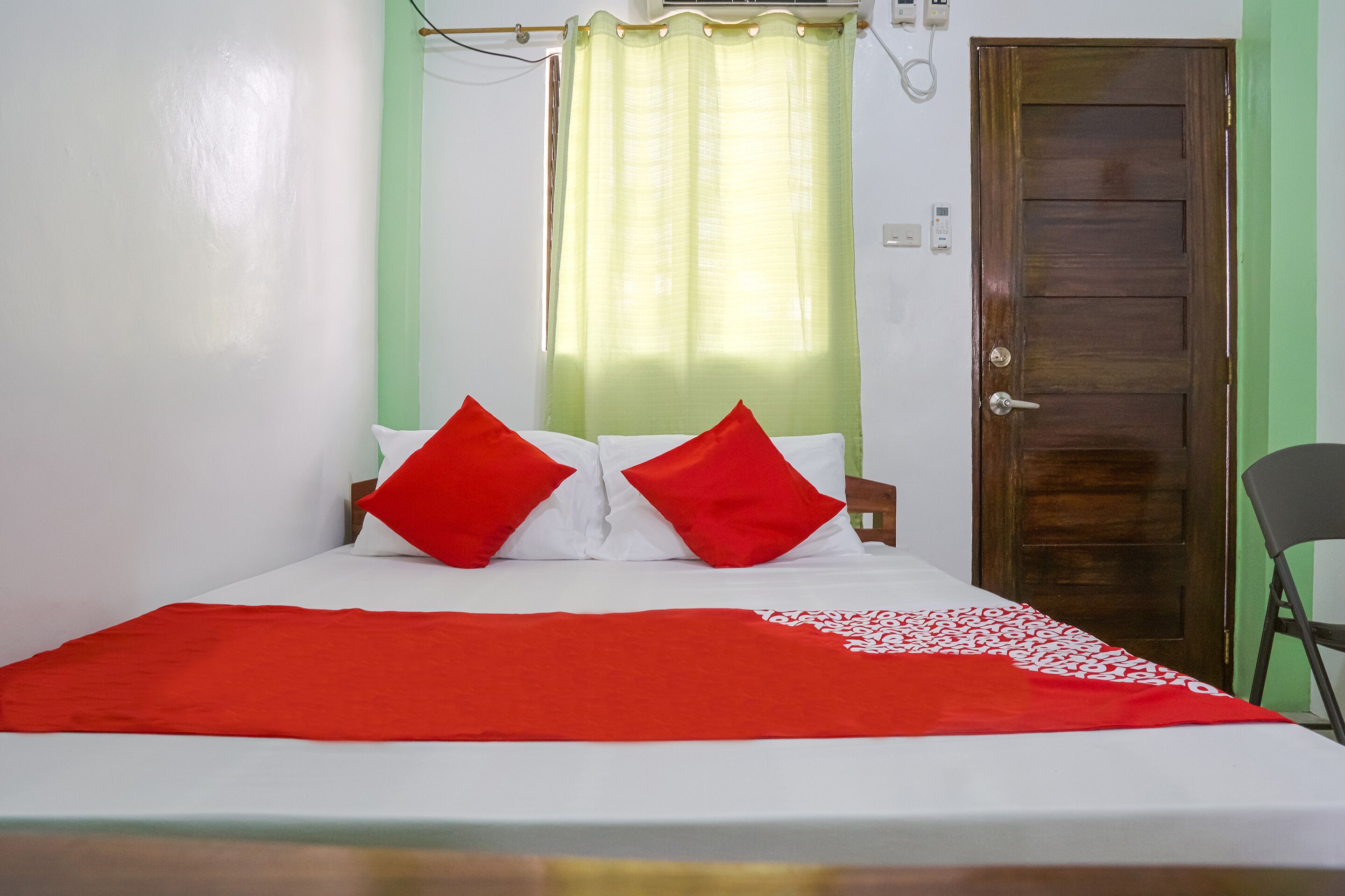 A bed with white bedding and red pillows, a red throw blanket, and a green curtain.