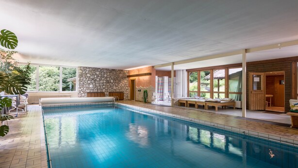 Une piscine intérieure avec des murs en pierre, des sièges en bois sur une terrasse et de grandes fenêtres.