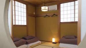 Una habitación tradicional japonesa con suelo de tatami, dos camas con ropa de cama morada, una lámpara colgante y un equipo de aire acondicionado en la pared.
