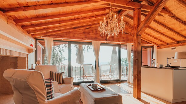 A spacious room with a wooden ceiling, a chandelier, a sofa, a dining table with a tray, and a view of the outdoors through large windows. - The Lake House (Orhangazi)