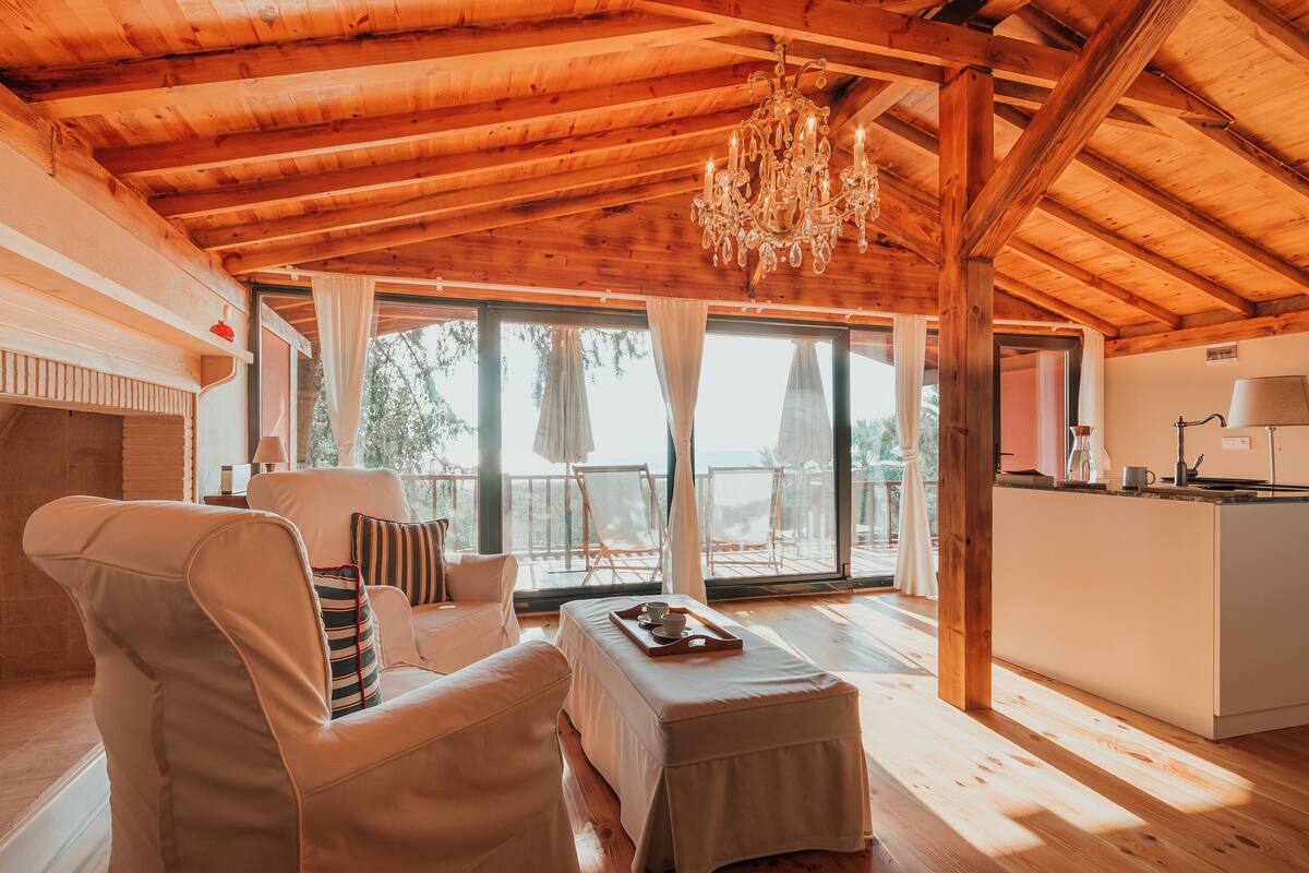 A spacious room with a wooden ceiling, a chandelier, a sofa, a dining table with a tray, and a view of the outdoors through large windows.