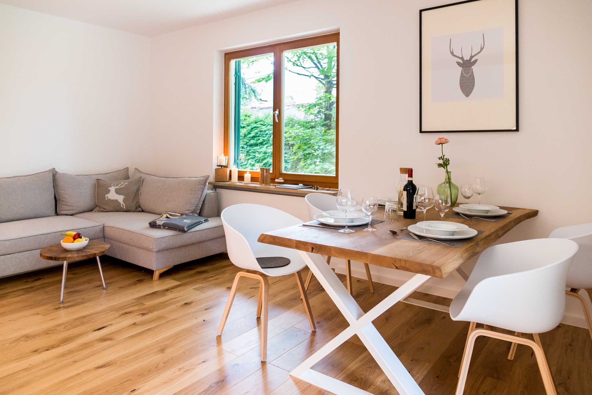 A modern living room with a wooden dining table, white chairs, a grey sofa, and a window with a view of greenery.