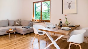 Un salón moderno con una mesa de comedor de madera, sillas blancas, un sofá gris y un ventanal con vistas a zonas verdes.