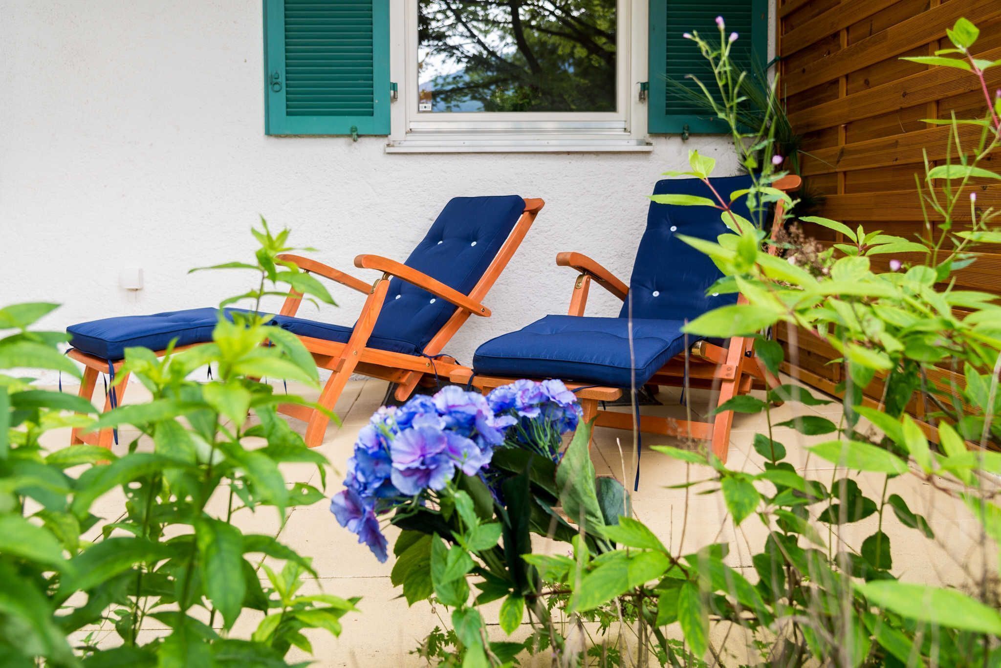 Zwei blaue Lounge-Stühle mit Holzgestellen und Kissen, vor einem weißen Gebäude mit grünen Fensterläden platziert.
