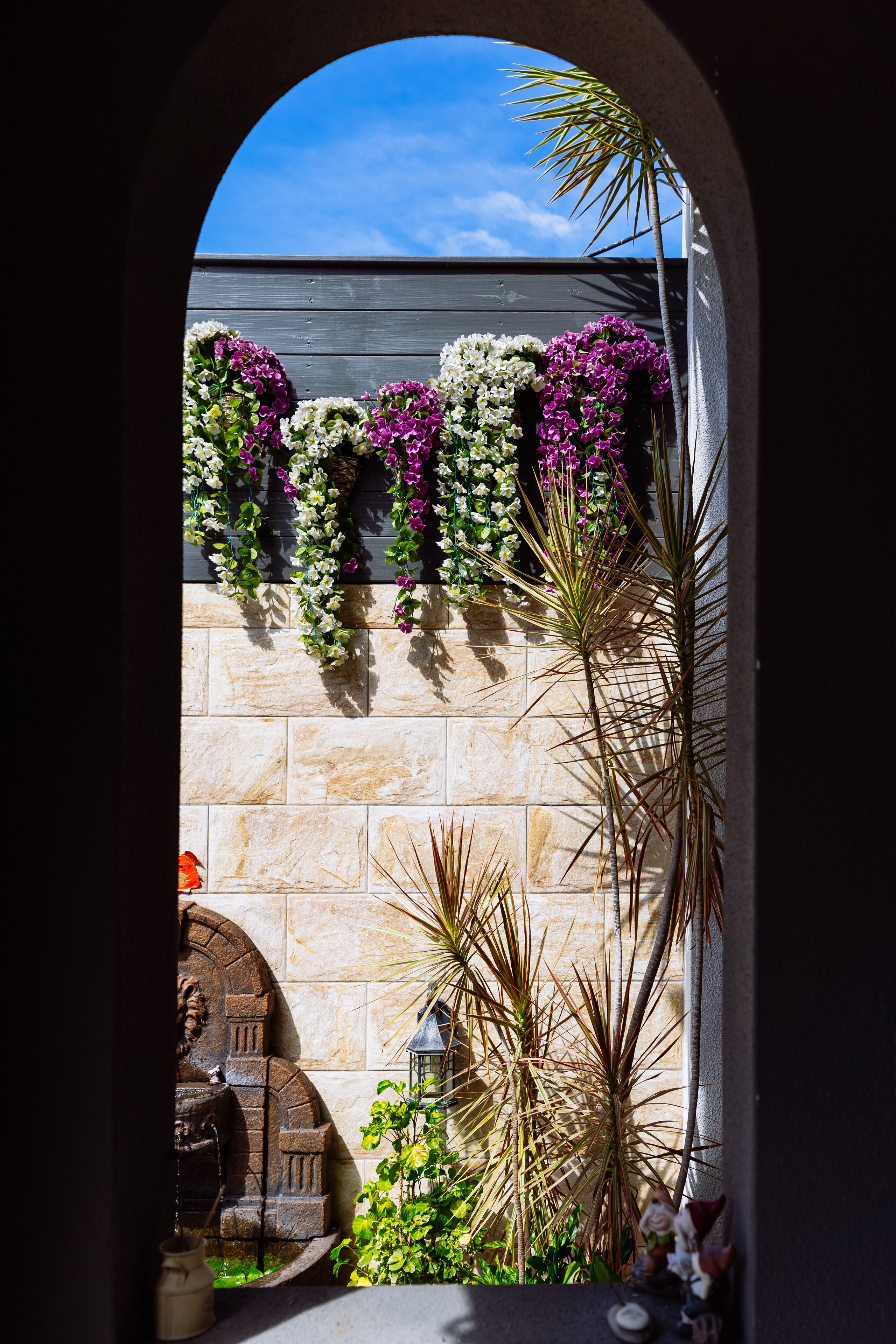 An arched doorway leading to a stone wall adorned with hanging flowers and plants, with a statue and a small fountain visible in the foreground.