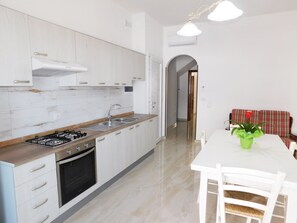 Een moderne keuken met witte kastjes, een gasfornuis en een spoelbak. Er is een eethoek met een tafel en stoelen, en een rode bloevas.