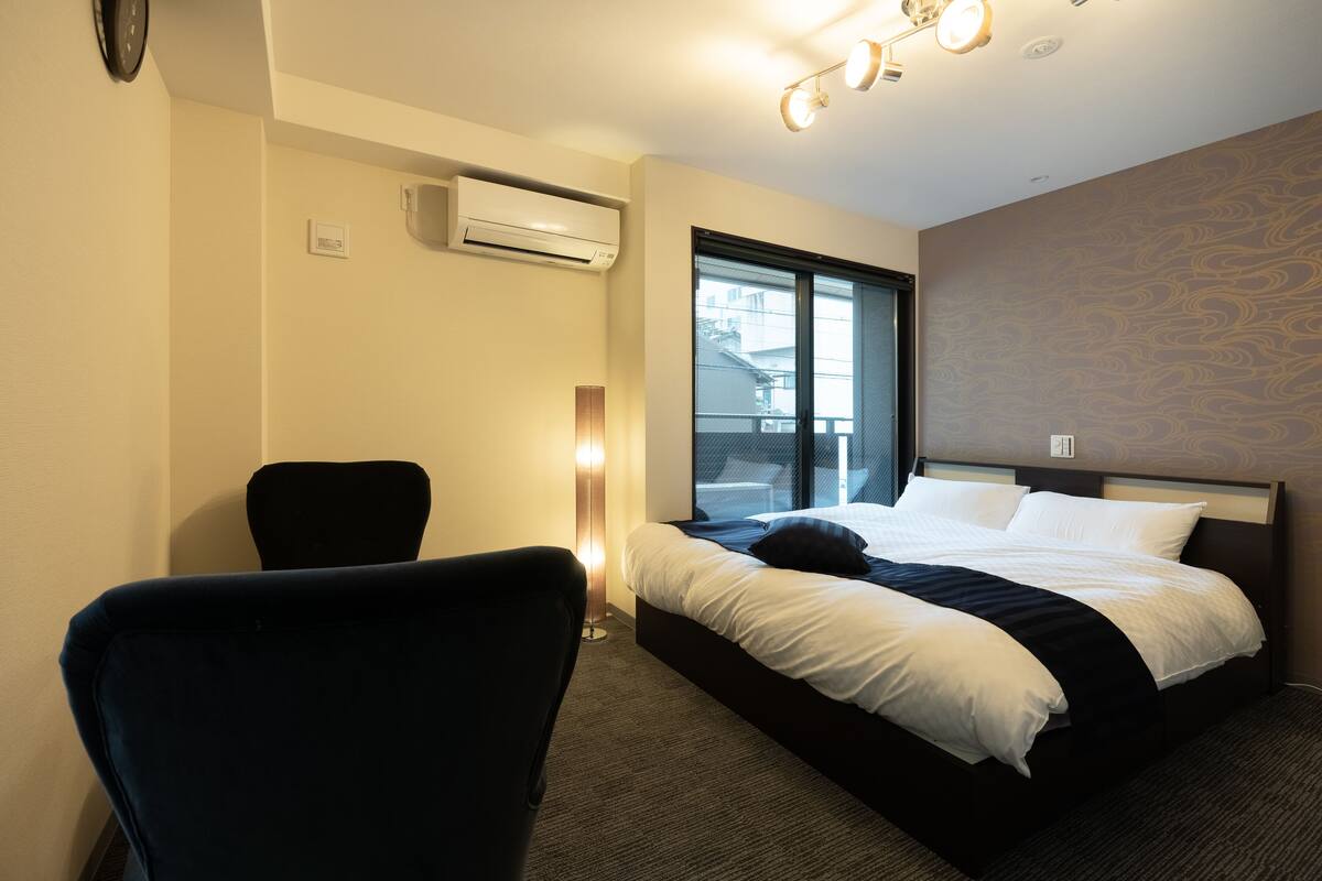 A hotel room with a bed, a chair, an air conditioning unit, a window, and a wall-mounted light fixture.