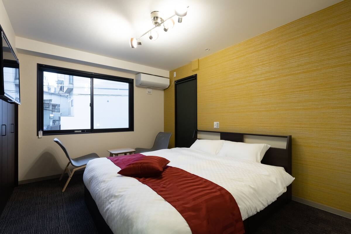 A hotel room with a bed, a desk, a chair, a window, and an air conditioning unit.