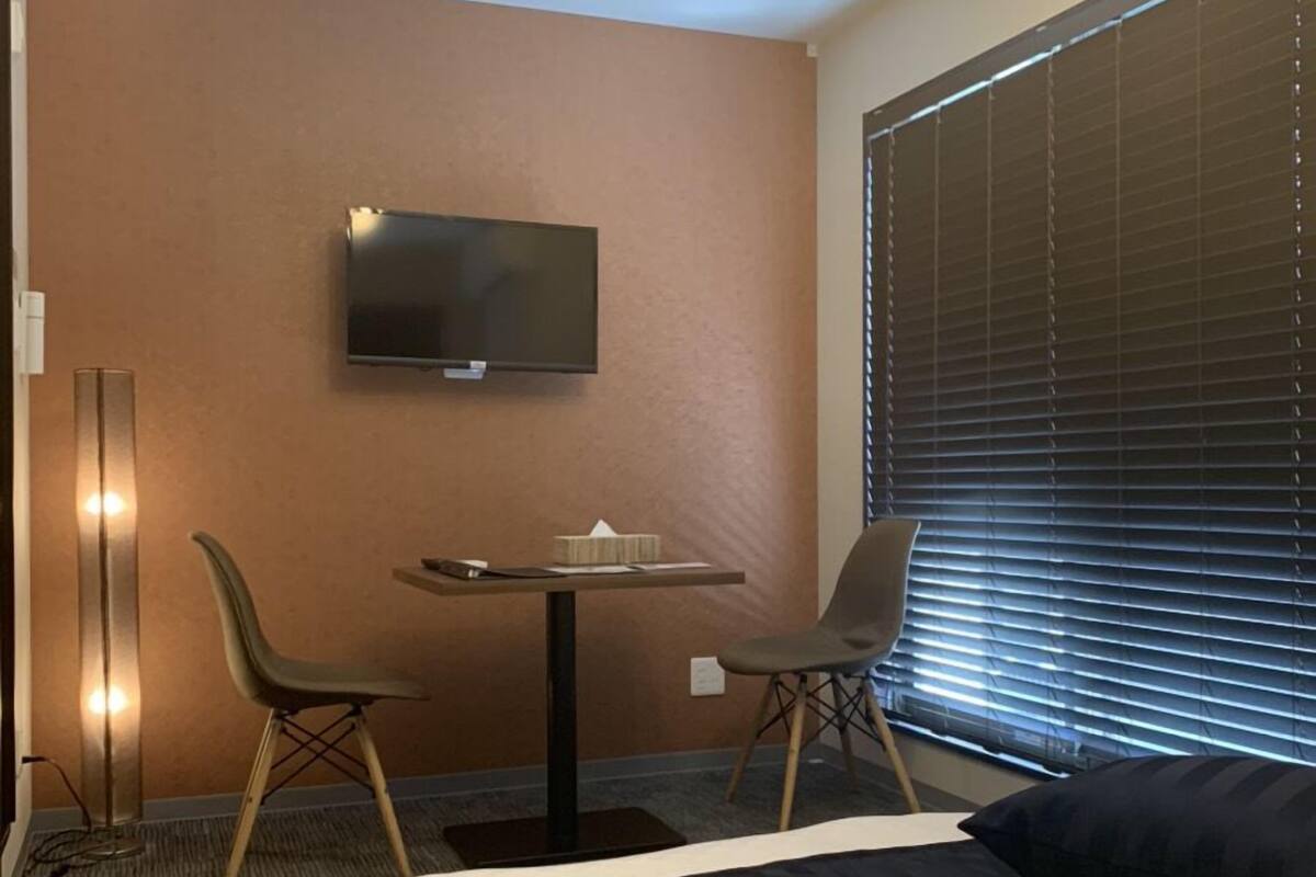 A hotel room with a bed, a desk with two chairs, a television on the wall, and a window with blinds.