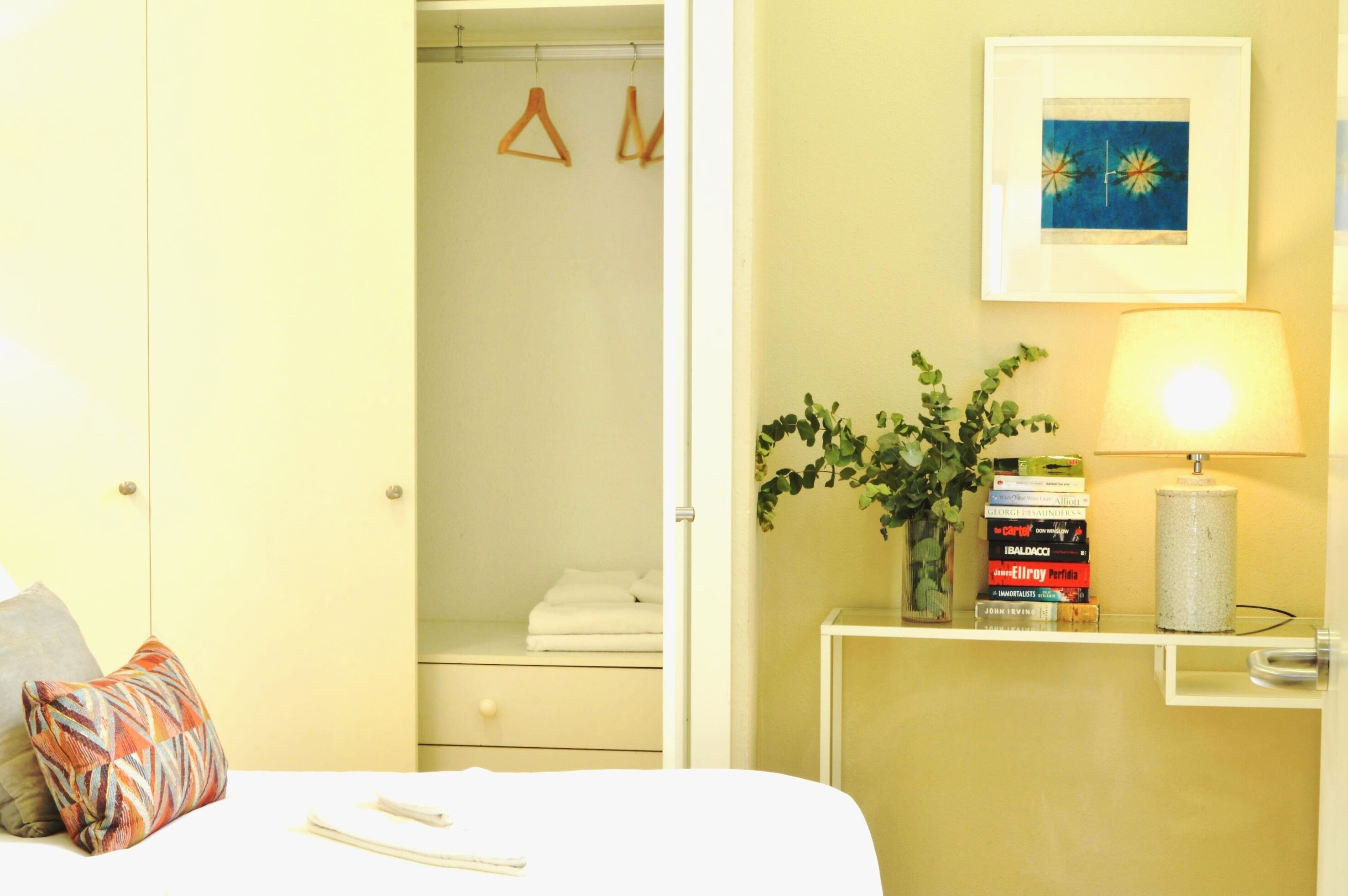 Une chambre d’hôtel avec un lit, une table de chevet avec une lampe et des livres, une armoire avec des cintres et un tableau encadré au mur.