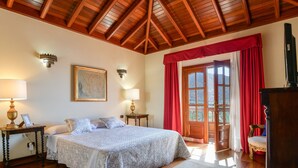 A bedroom with a wooden ceiling, a bed with a patterned bedspread, two bedside lamps, a nightstand, a chair, and a view through a window with red curtains. - Beautiful Villa La Petit with Pool, Terrace, Garden and Wi-Fi (GUATIZA)