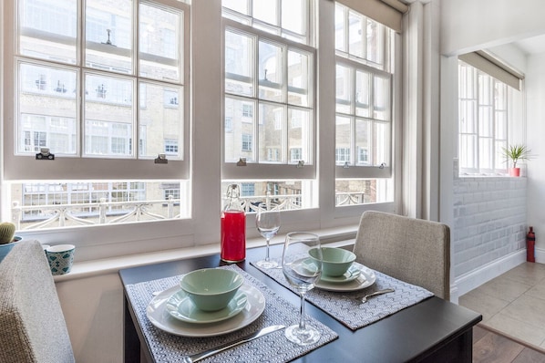 Un comedor con una mesa preparada para dos, ventanas amplias con vista a edificios, una planta en maceta sobre una estantería y un extintor en la pared.