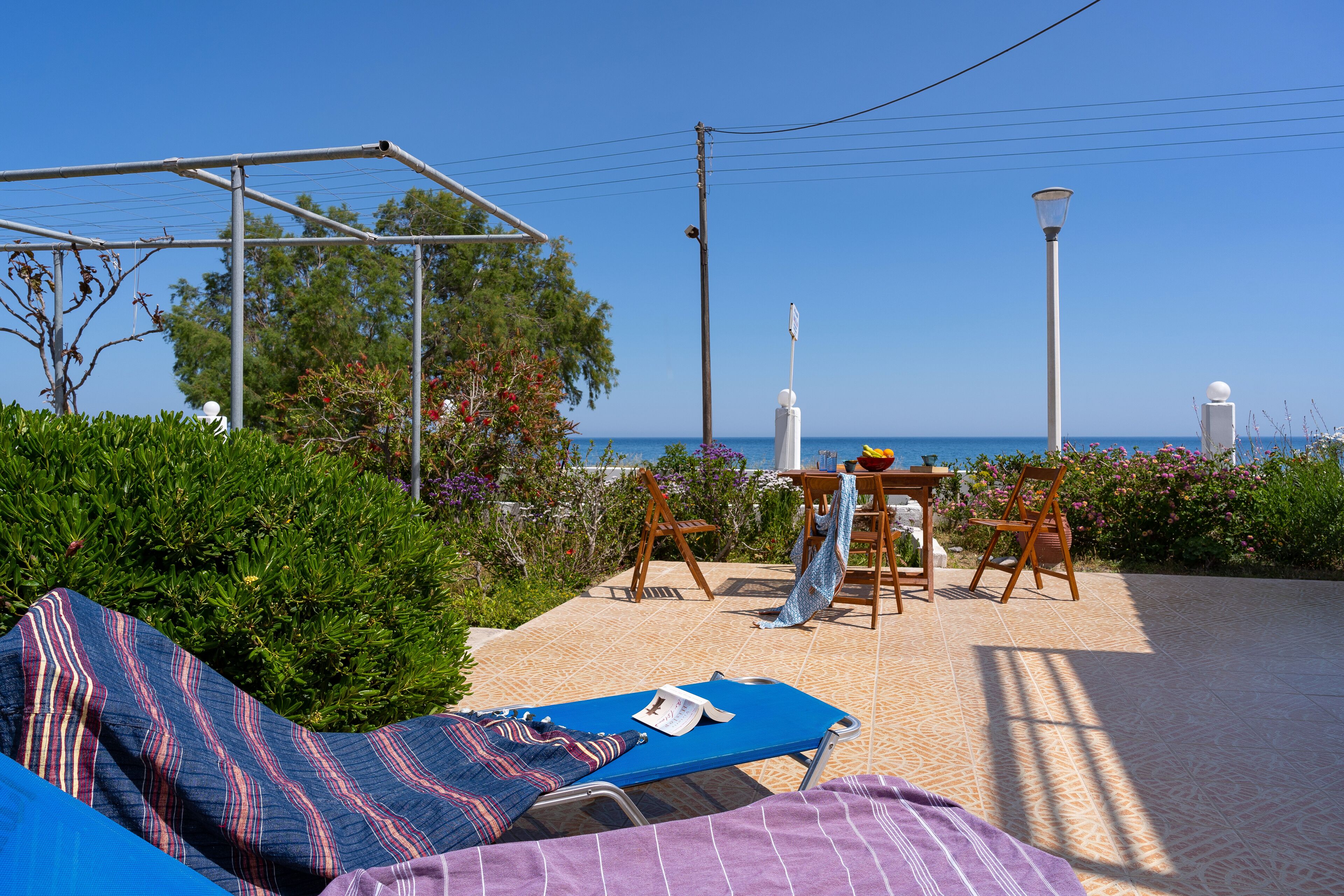 Ein sonniger Außenbereich mit Lounge-Sitzgelegenheit, einem kleinen Tisch und einem Stuhl, umgeben von viel Grün und mit Blick aufs Meer.