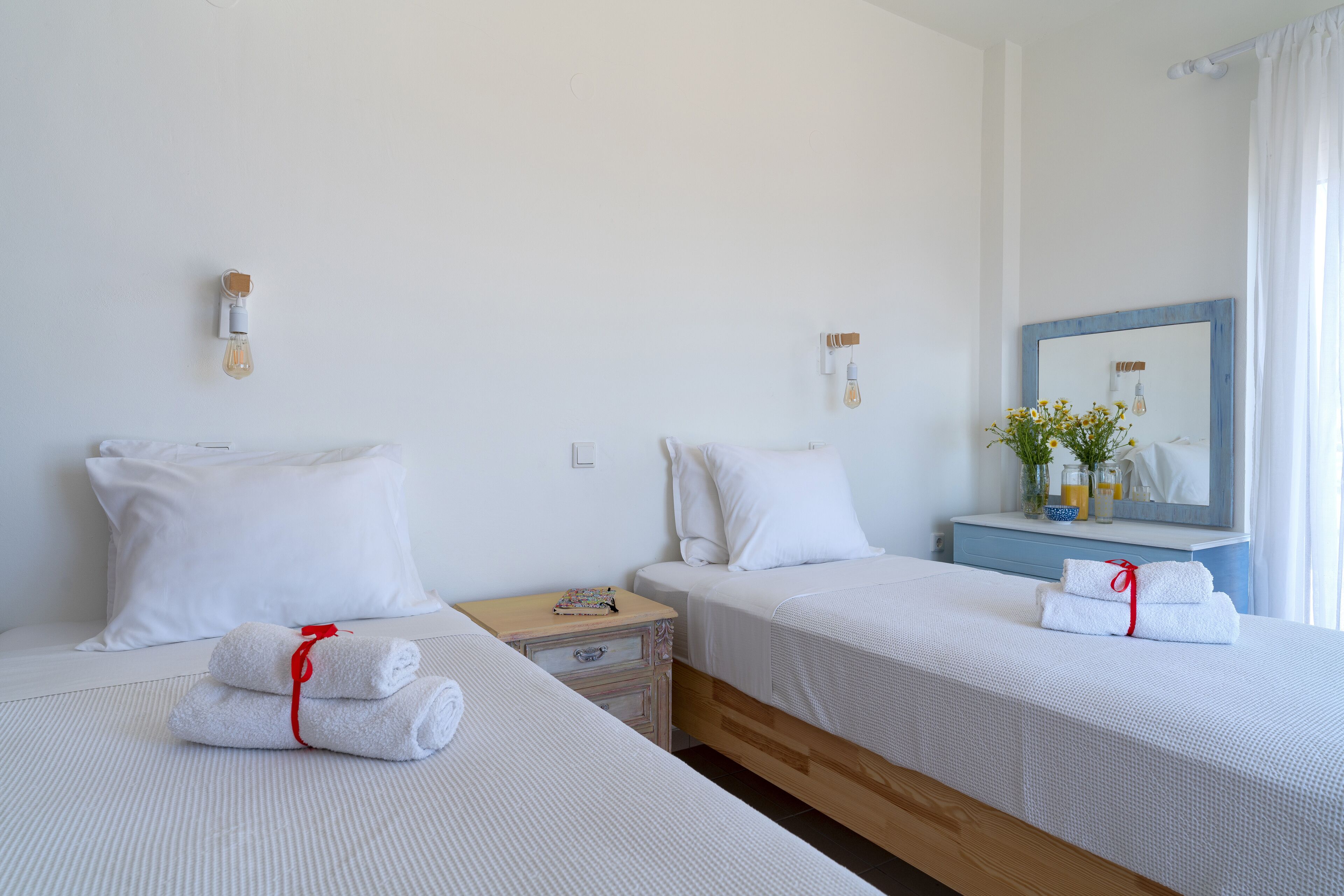 Une chambre d’hôtel avec deux lits, chacun recouvert d’une literie blanche et agrémenté d’une serviette rouge nouée en forme de ruban. On y trouve une table de chevet en bois avec une lampe, ainsi qu’une petite table avec un livre. Un miroir à cadre bleu et un vase rempli de fleurs sont posés sur la commode.