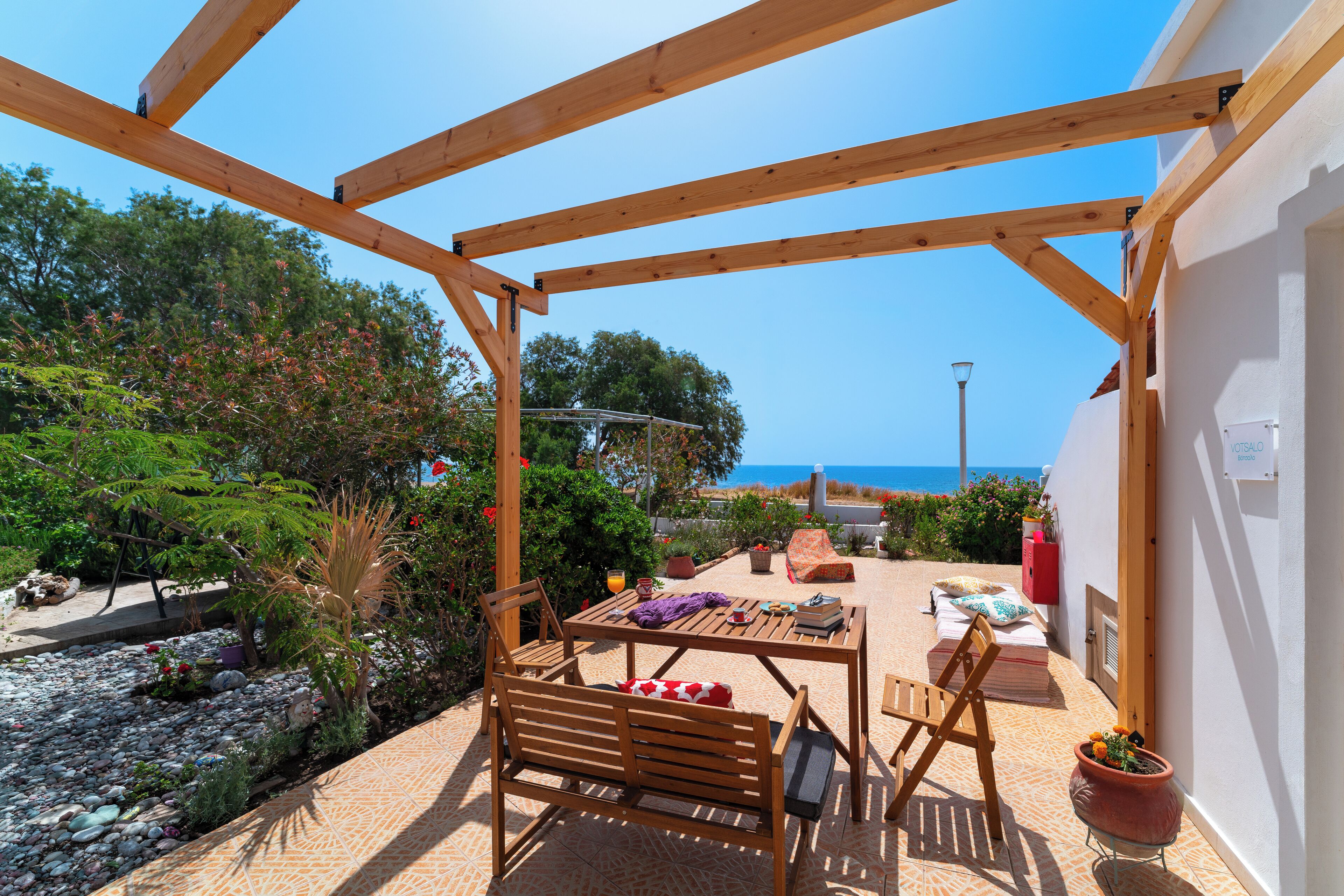 A wooden pergola patio with a table and chairs, surrounded by potted plants and a view of the sea.