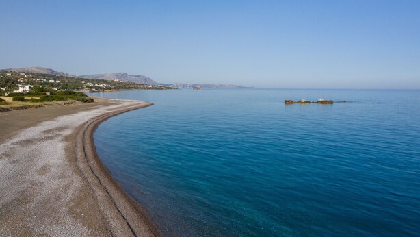 A coastal landscape with a clear blue sea, a sandy beach, and a few buildings in the distance. - Sea & Sun (Rhodes)