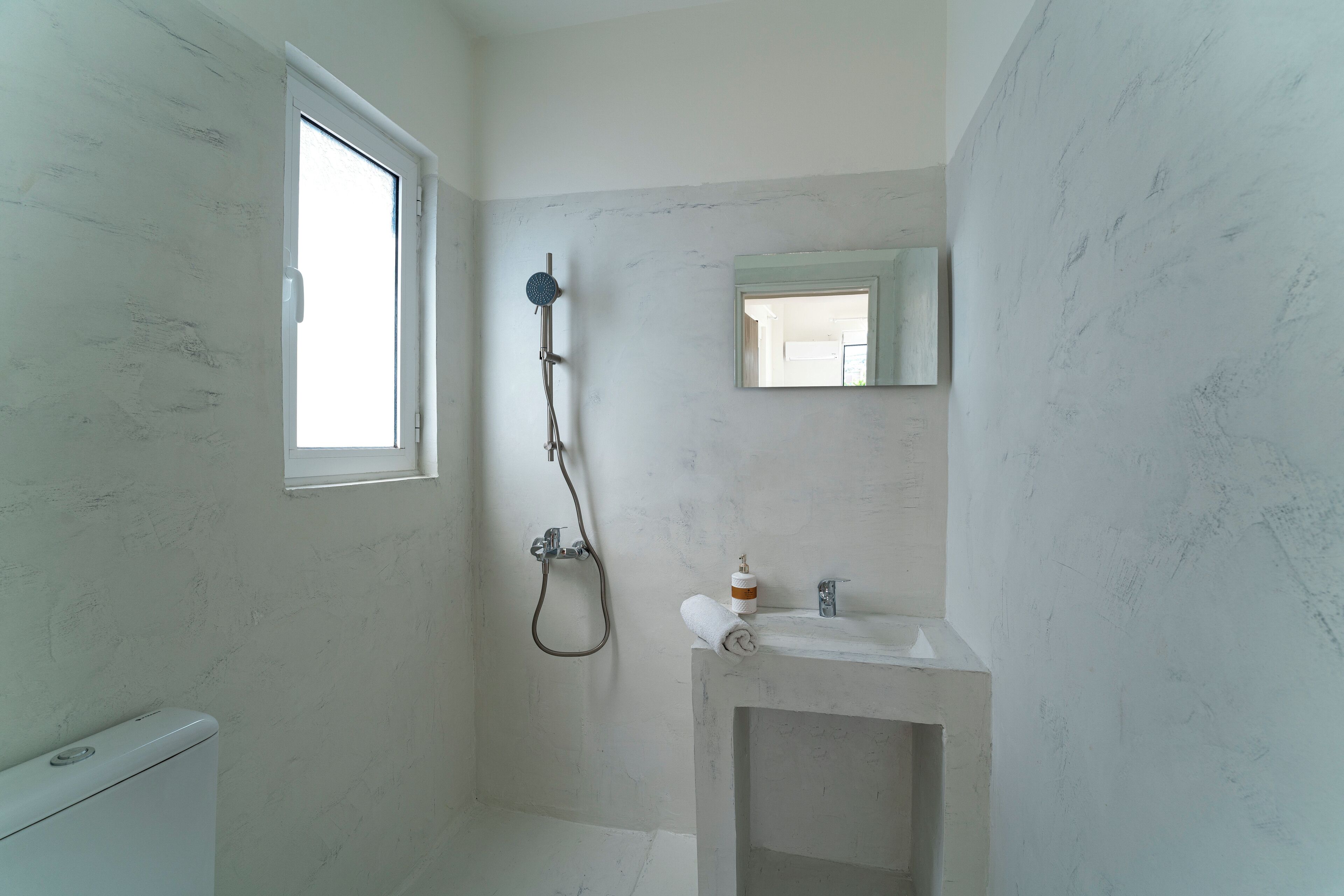 A bathroom with a toilet, a shower with a handheld showerhead, a sink with a mirror, and a window.