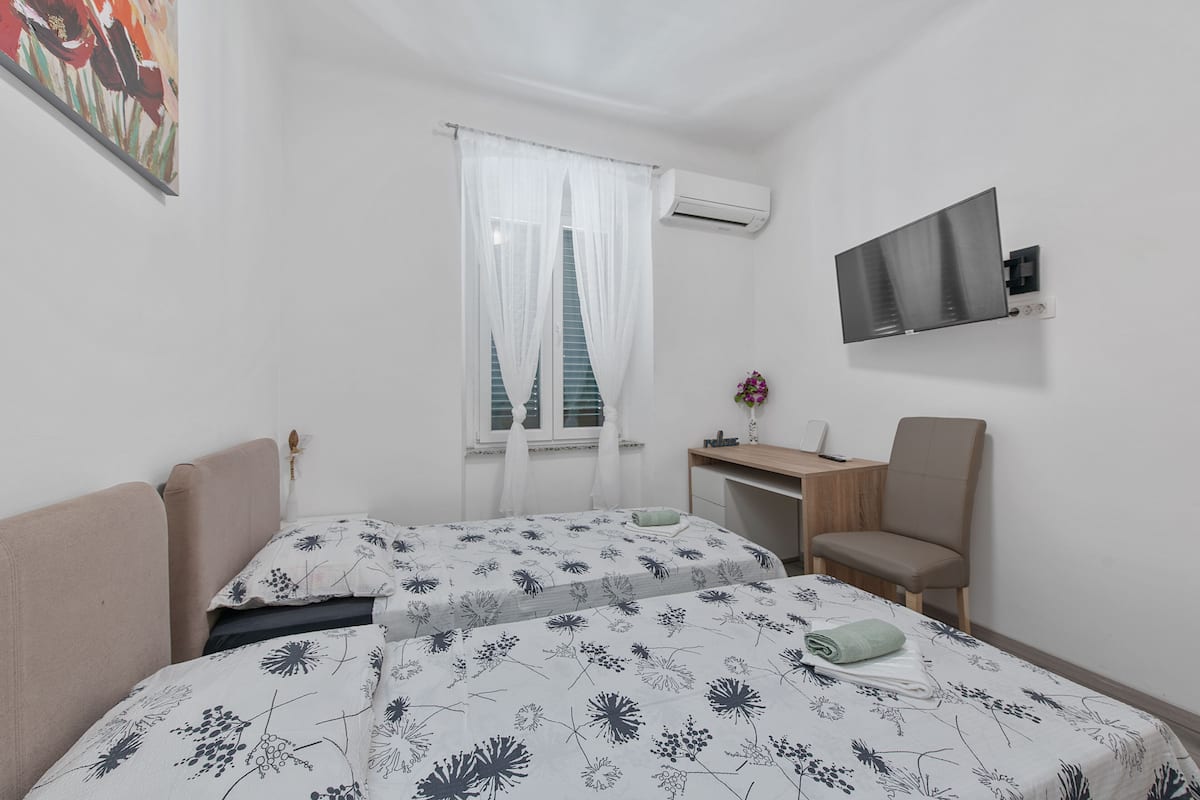 A hotel room with two beds, a desk, a chair, an air conditioner, and a TV mounted on the wall.