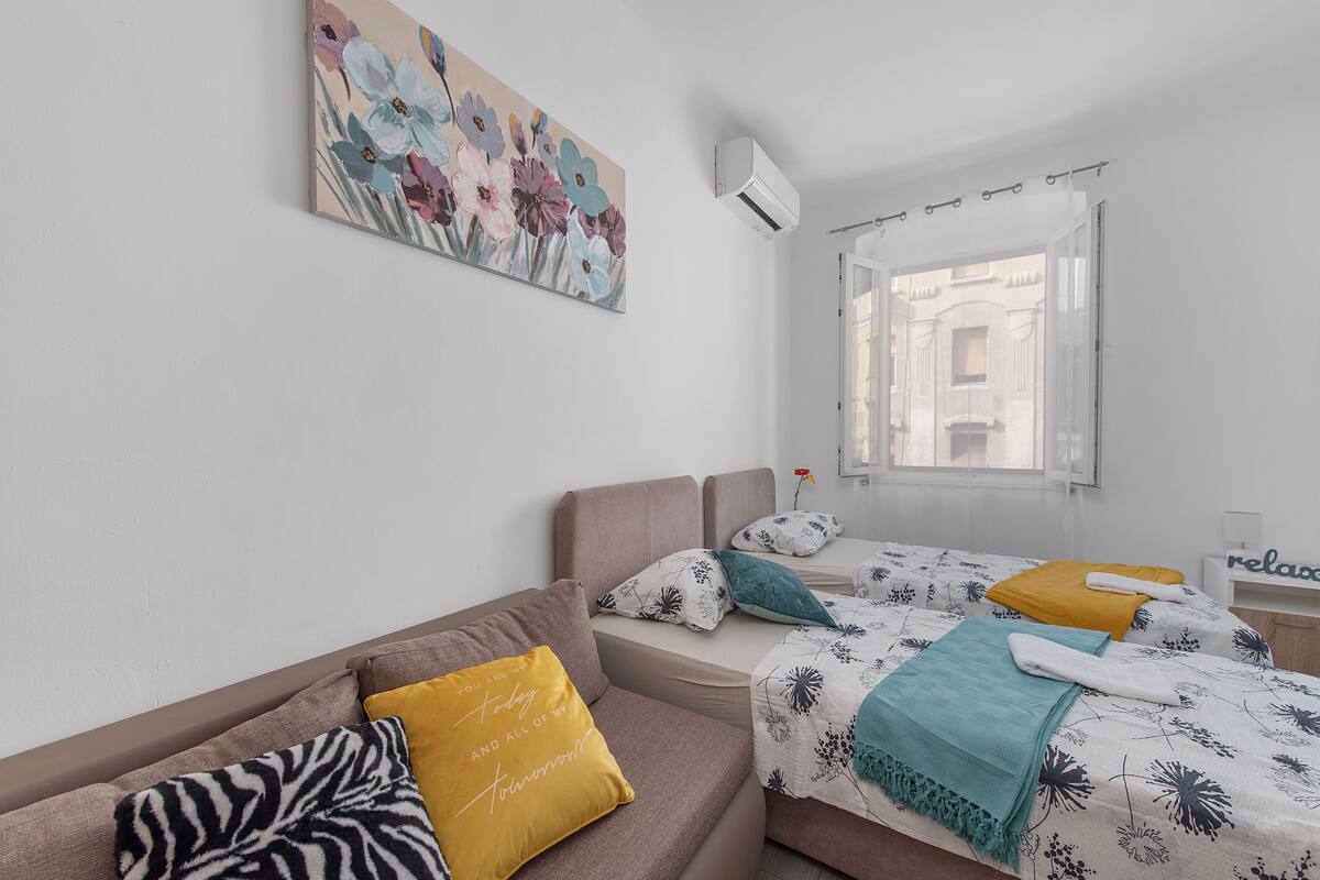 A modern hotel room with a sofa, two beds with floral bedding, a zebra-patterned pillow, and a large floral painting on the wall.