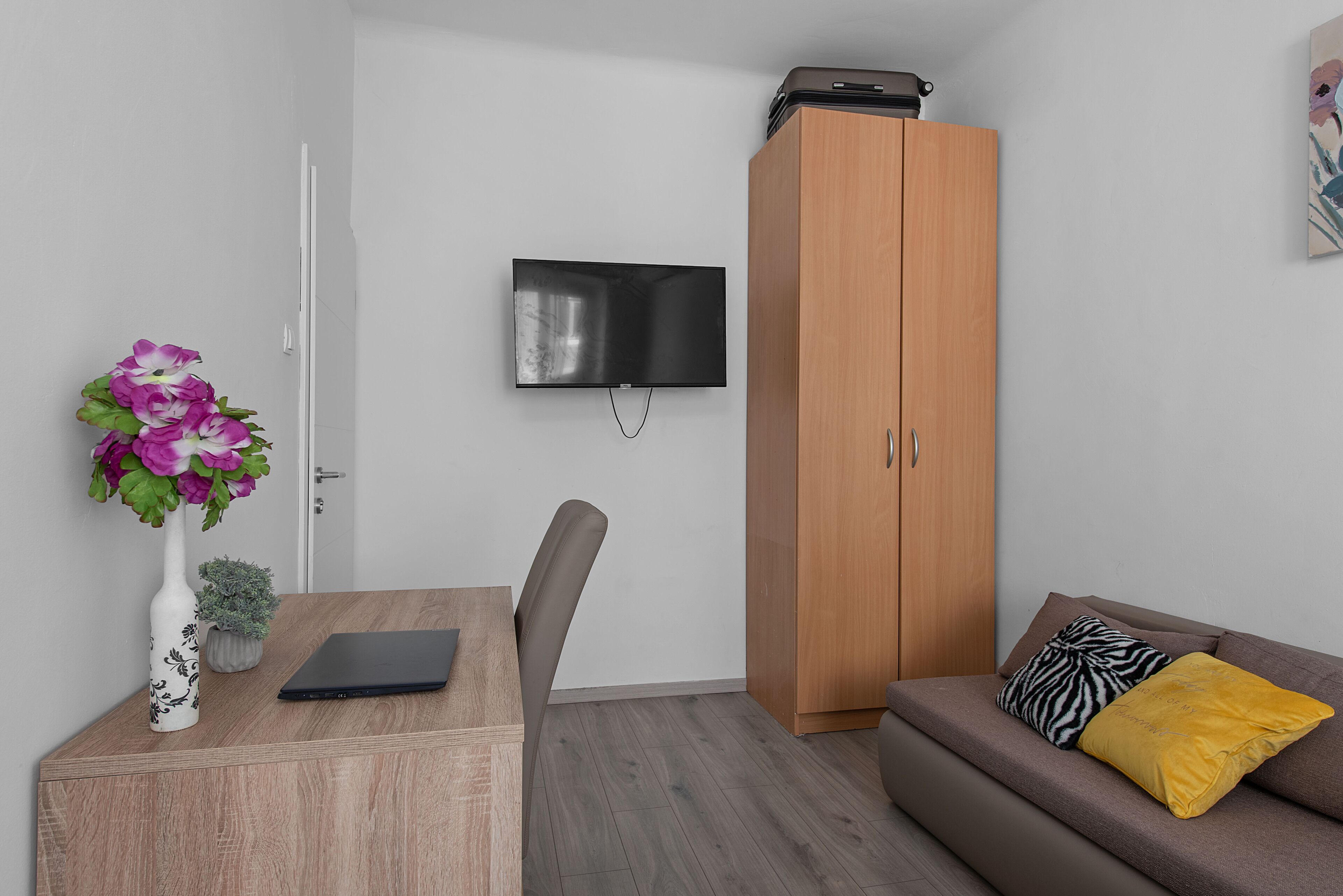 Quarto moderno com escrivaninha de madeira, um laptop, televisão fixada na parede, sofá marrom com almofadas e guarda-roupa de madeira.