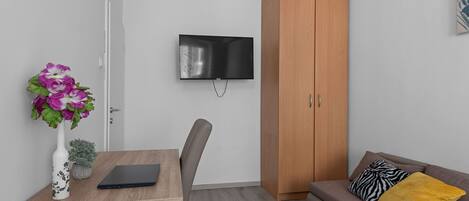 Quarto moderno com escrivaninha de madeira, um laptop, televisão fixada na parede, sofá marrom com almofadas e guarda-roupa de madeira.