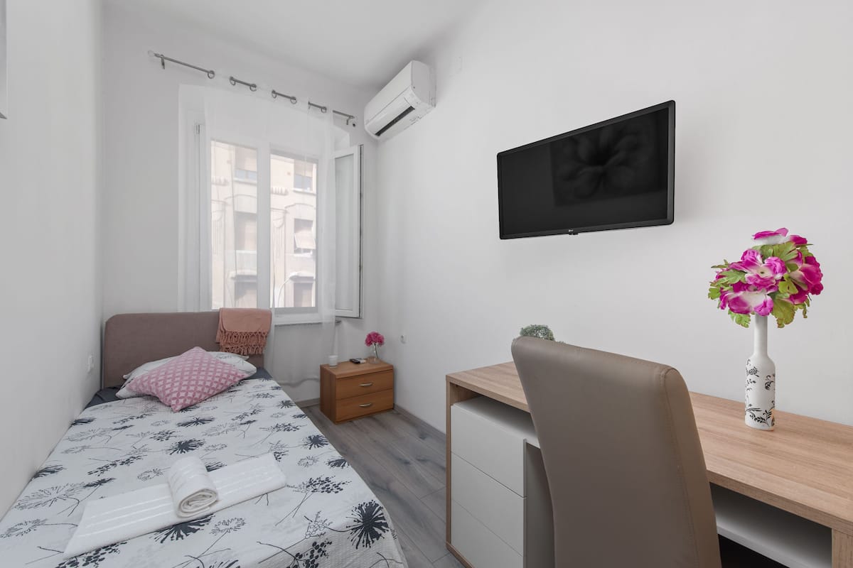 A bedroom with a bed, a desk, a chair, a TV, a nightstand, and a vase of flowers.