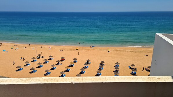 A beach with sun loungers and umbrellas, a clear blue sea, and a white building in the foreground.