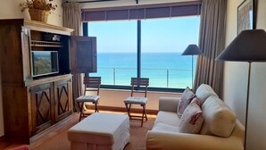 A living room with a sofa, two chairs, a TV, and a view of the sea. - Albufeira Beach Cliffs House by Rentals in Algarve (Albufeira)