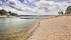 Une plage de galets avec une jetée s’avançant dans une eau calme, entourée d’arbres et surplombée d’un ciel nuageux.