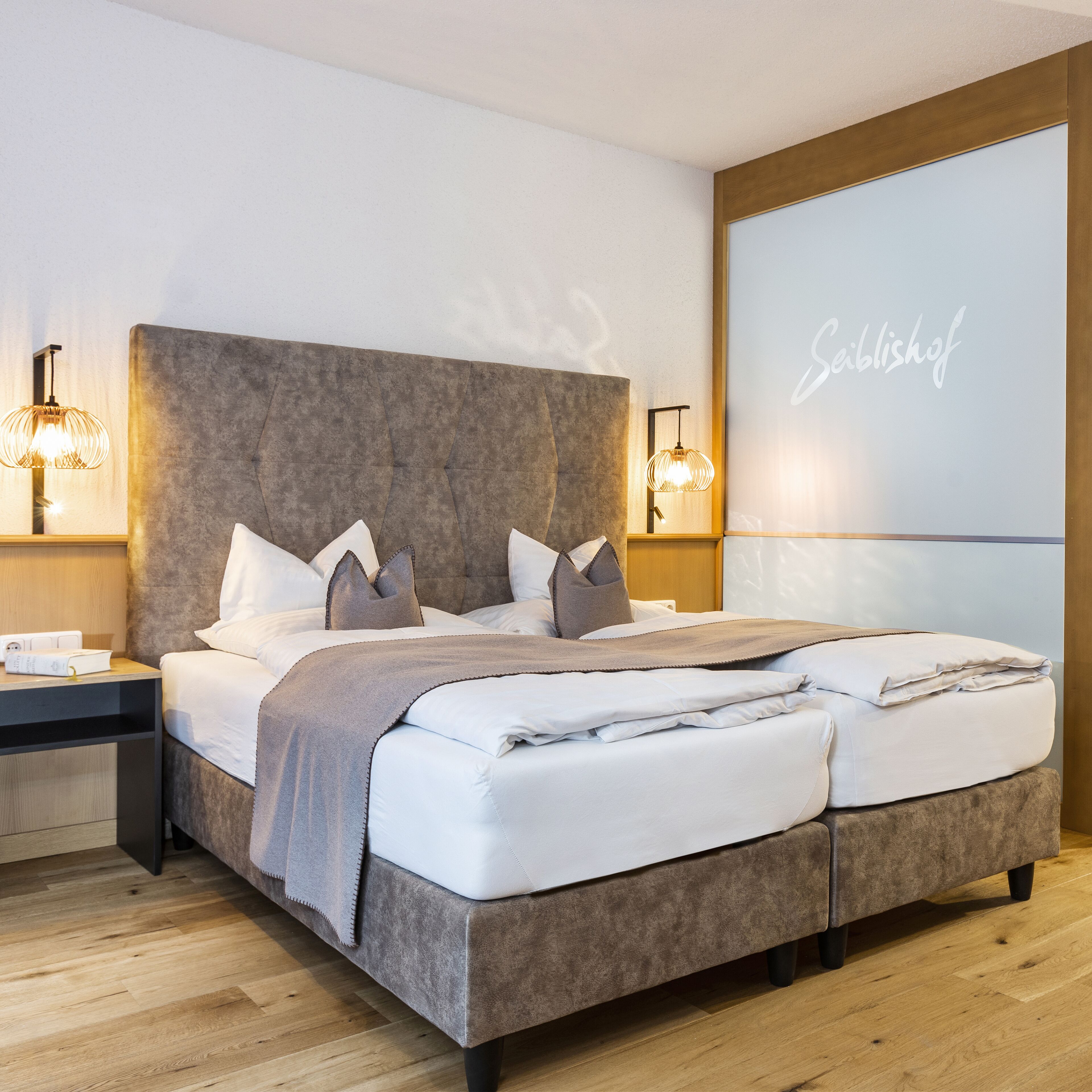 A modern bedroom with a large bed, two bedside tables, and a wall-mounted projector screen displaying "Seiblshof".