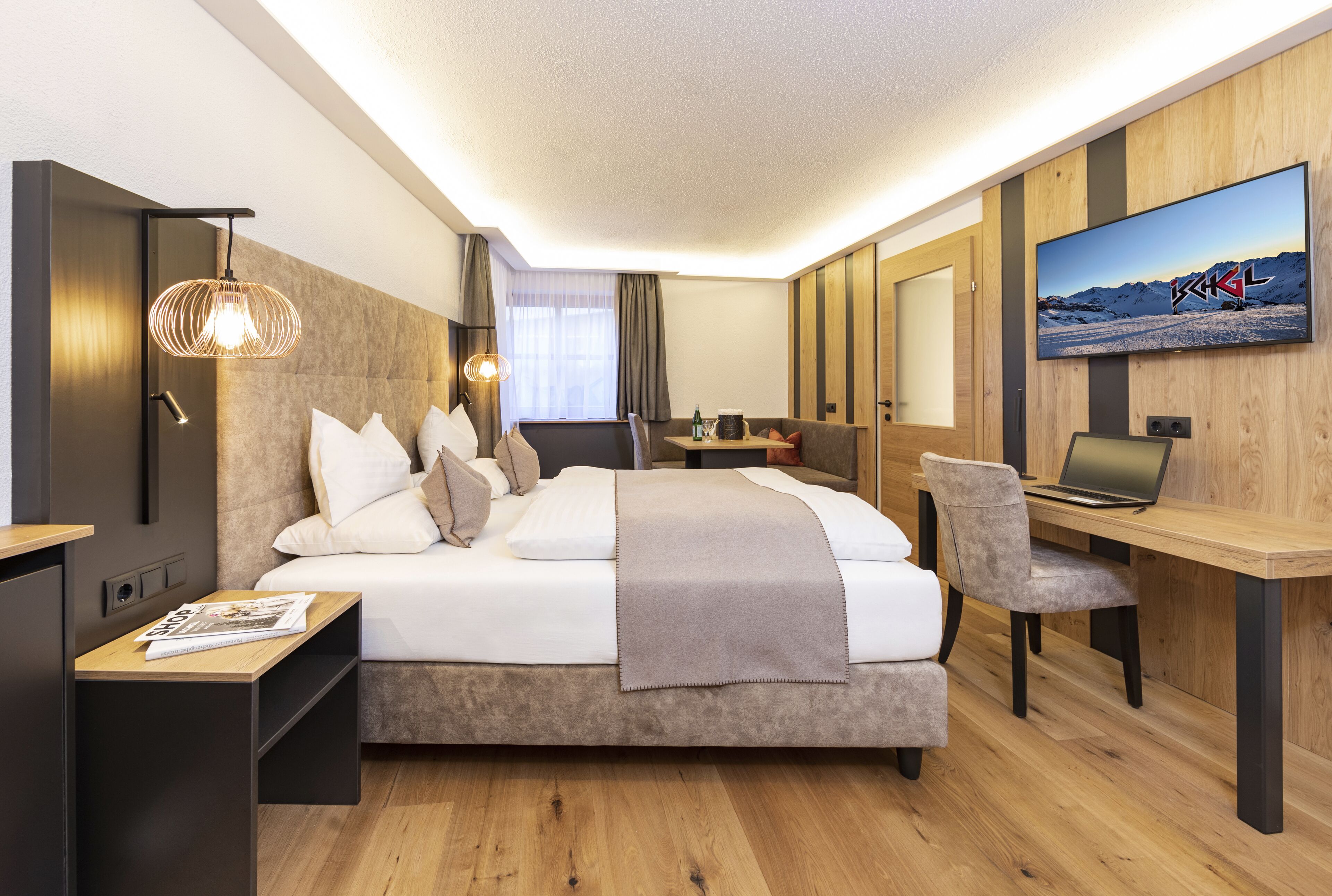 A modern hotel room with a large bed, a desk with a laptop, a TV mounted on the wall, and a wooden floor.