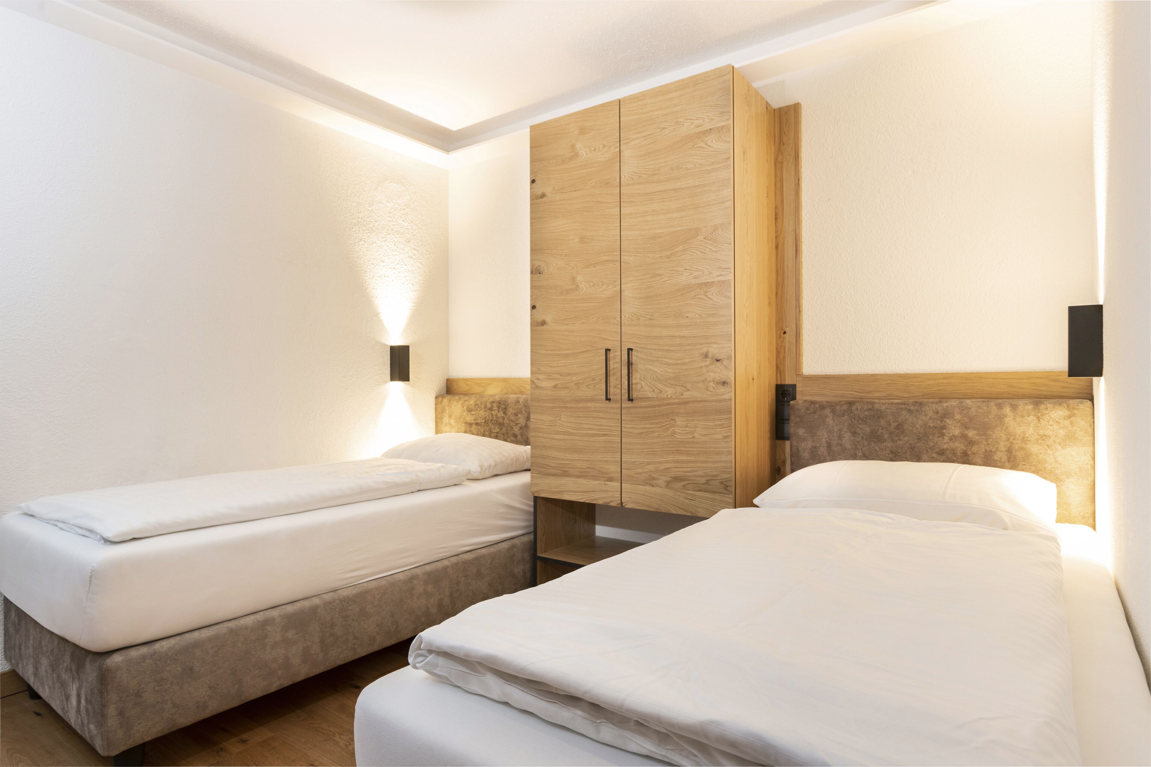 A room with two single beds, a wooden wardrobe, and a nightstand with a lamp.