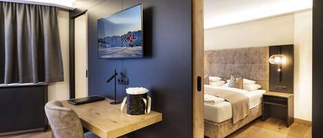 A modern hotel room with a wooden desk, a bed with a headboard, a mounted TV, and a bedside table with a lamp.