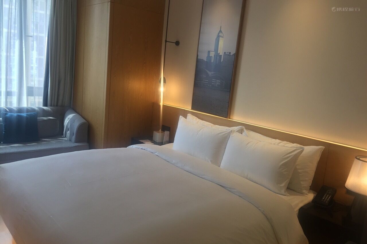 A hotel room with a large bed, a nightstand with a lamp, a window with curtains, and a cityscape painting on the wall.