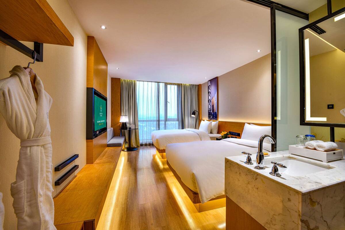 A hotel room with two beds, a large window, a TV, a desk, and a bathroom with a sink and mirror.