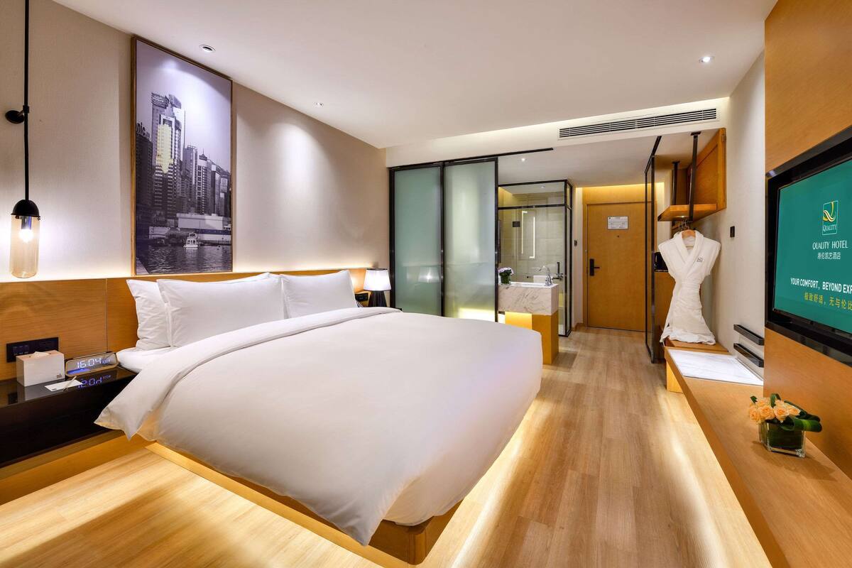 A modern hotel room with a large bed, a nightstand, a digital clock, a framed cityscape picture, a sliding glass door to the bathroom, and a television displaying the hotel name.