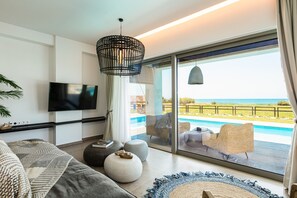 A modern living room with a large TV, comfortable seating, and a view of a pool and ocean. - Feyre Boutique Mansion-Echoes of the Sea by etouri (Rethymno)