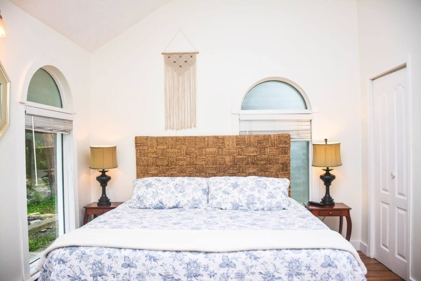 A bedroom with a woven headboard, floral bedding, two bedside lamps, and a view of greenery through a window.