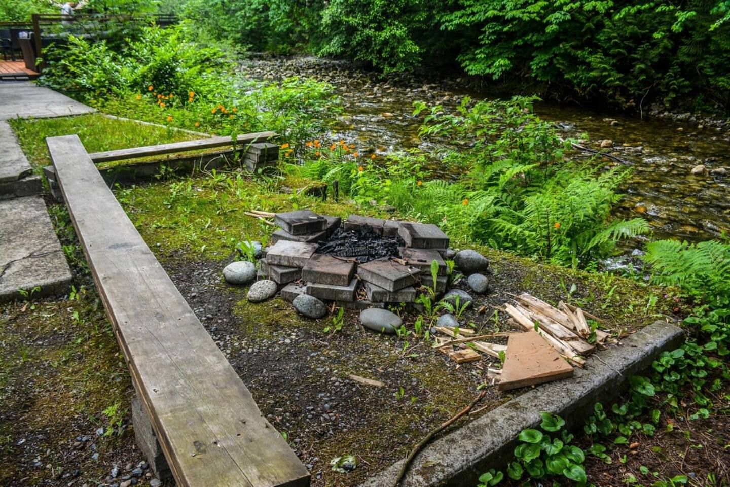 A natural outdoor setting with a fire pit surrounded by rocks and logs, a wooden deck, and a stream with surrounding vegetation.