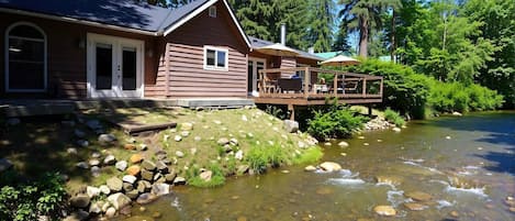 Une cabane dotée d’une terrasse donnant sur un ruisseau aux eaux claires, le tout entouré d’arbres.