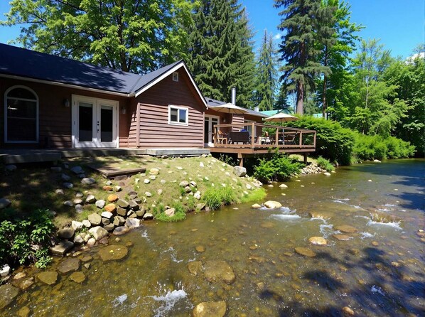 A cabin with a deck overlooking a clear stream, surrounded by trees.