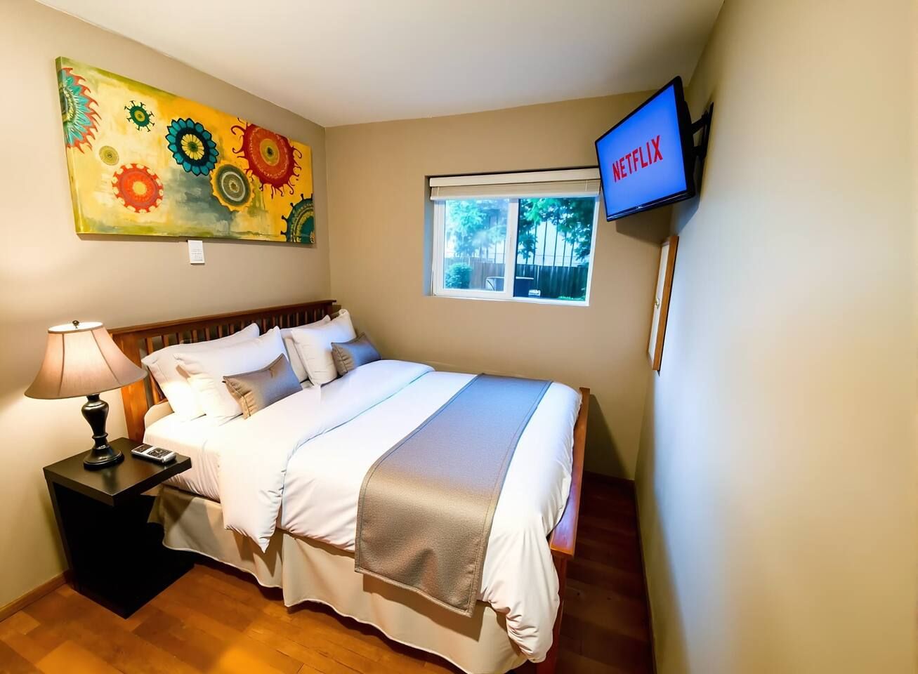 A hotel room with a bed, a nightstand, a lamp, a painting, a window, and a TV displaying Netflix.