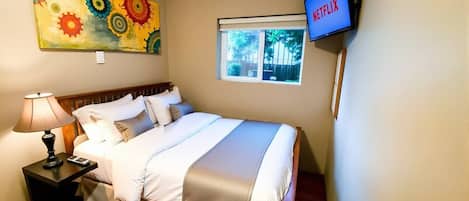 Habitación de hotel con cama, mesita de noche, lámpara, un cuadro, ventana y televisor mostrando Netflix.
