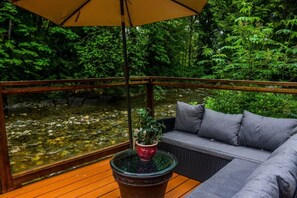 A wooden deck with a sofa and a table, overlooking a stream and dense greenery.