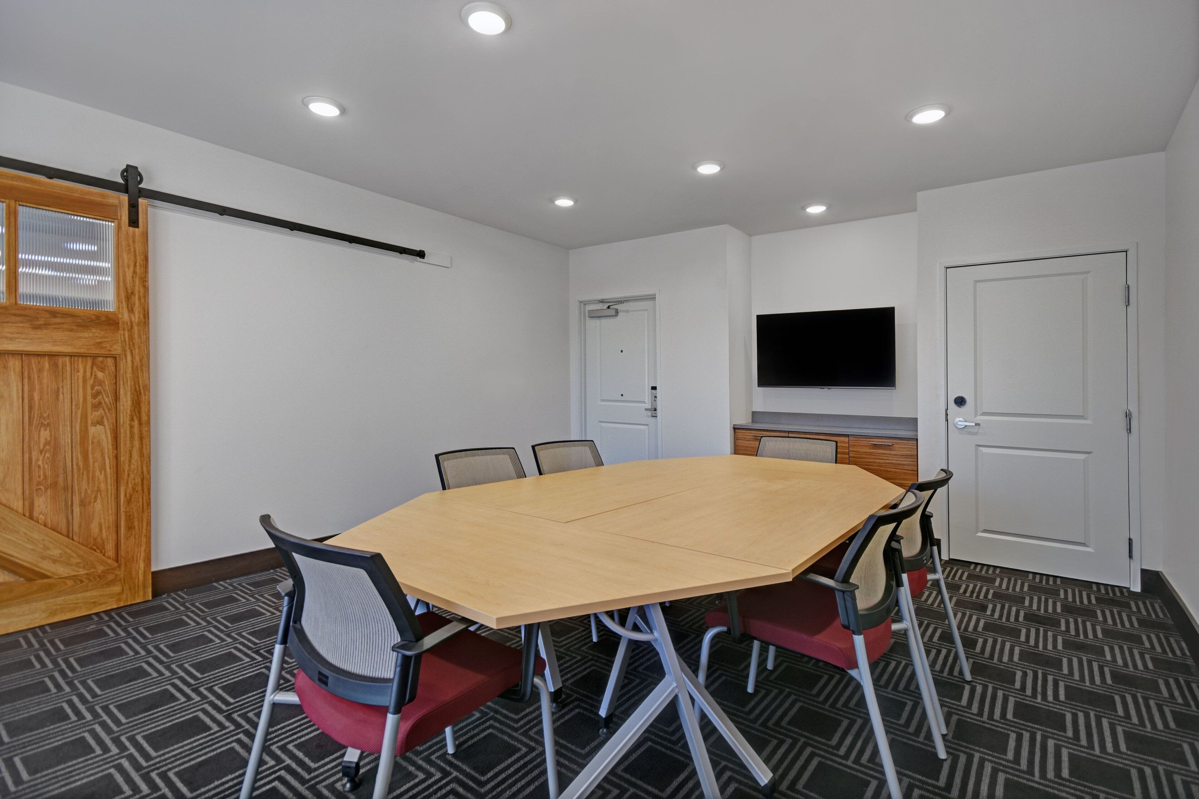 Une salle de conférence avec une grande table, six chaises, une porte en bois, une porte blanche, un téléviseur fixé au mur et un tapis à motifs.