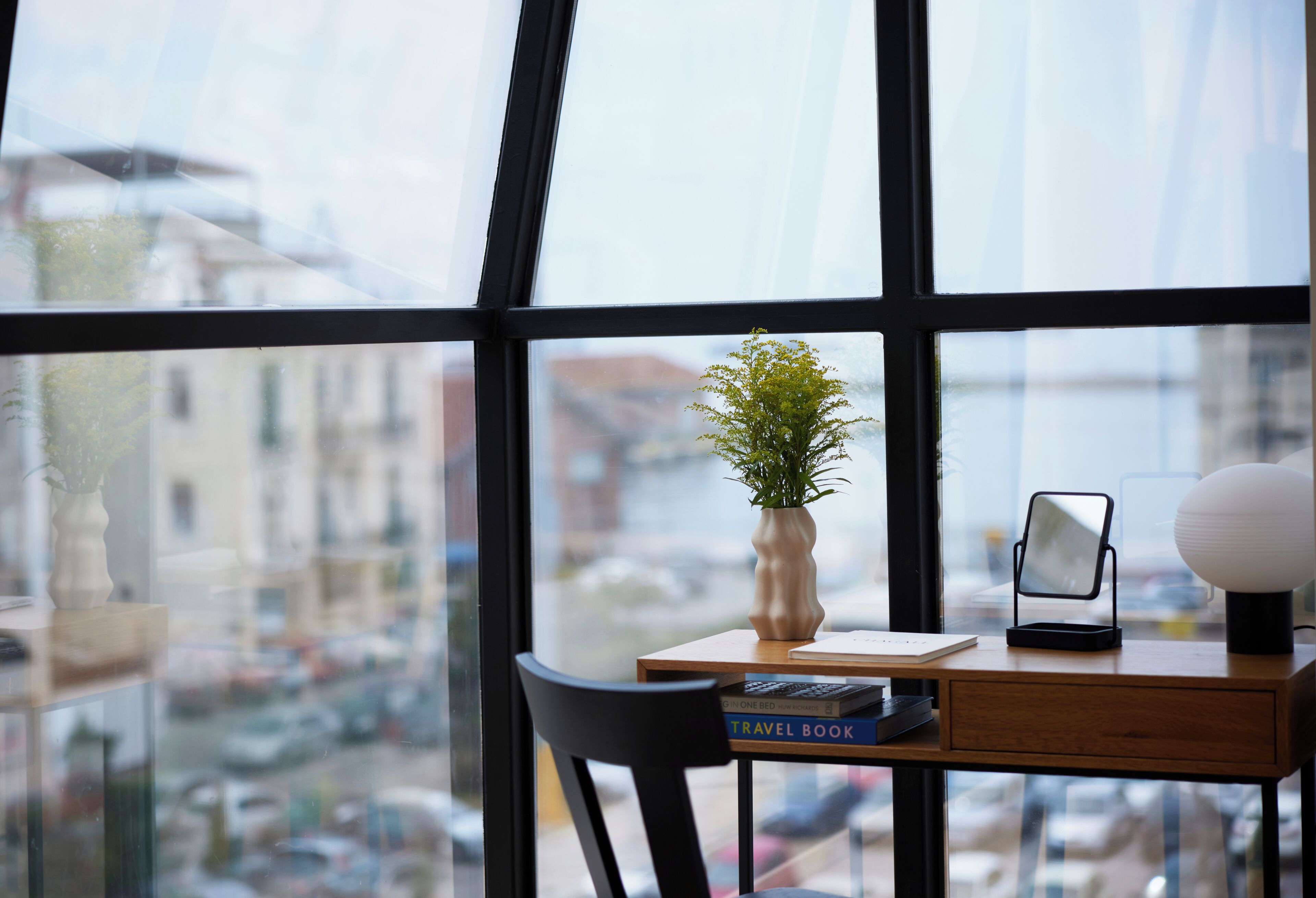 Blick durch ein Fenster auf einen Schreibtisch, eine Pflanze, ein Buch und eine Lampe.