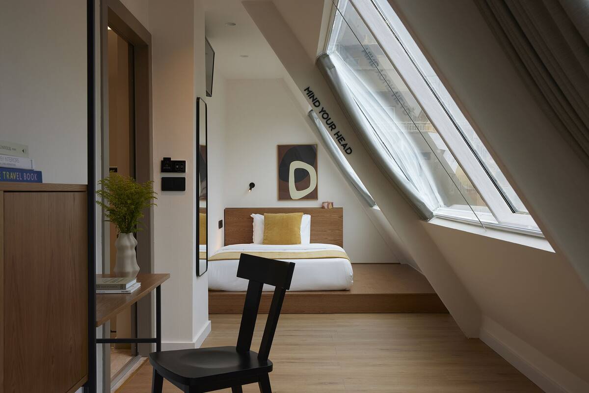 A modern bedroom with a skylight, a bed with white and mustard bedding, a wooden headboard, and a bedside table with a plant.